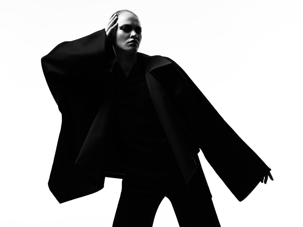 Editorial portrait in a structured black coat, frost light on a white wall